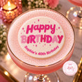 Load image into Gallery viewer, A pink frothy cocktail in a coupe glass with a gold rim, topped with a round white foam lid printed with 'Happy Birthday Sophie's 40th Birthday' in pink lettering and small star accents. The drink is photographed on a reflective tabletop with blurred pink candles and flowers in the background.