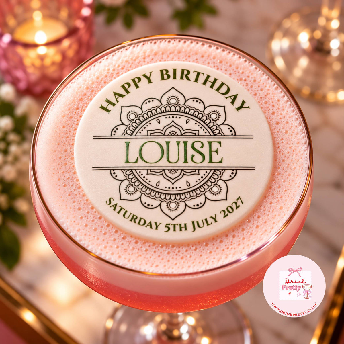 A pink frothy cocktail in a coupe glass with a gold rim, topped with a round white foam insert printed with 'HAPPY BIRTHDAY LOUISE' and a decorative mandala design. The glass is photographed on a softly lit table with candles and flowers in the background.