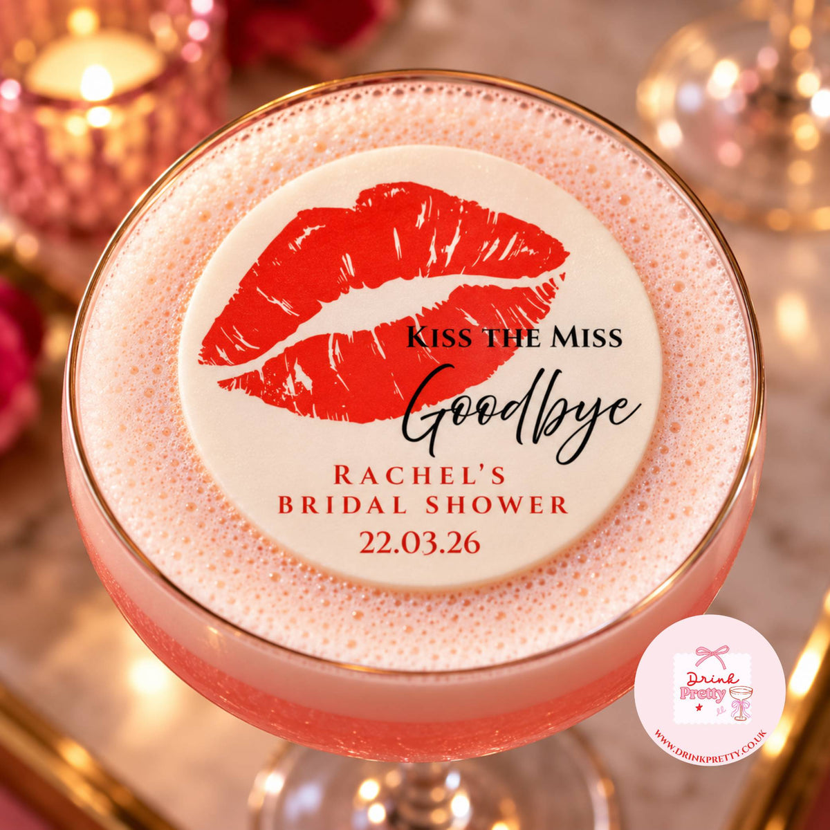 A pink frothy cocktail in a coupe glass with a round white topper featuring red lips and the text 'KISS THE MISS Goodbye Rachel's Bridal Shower 22.03.