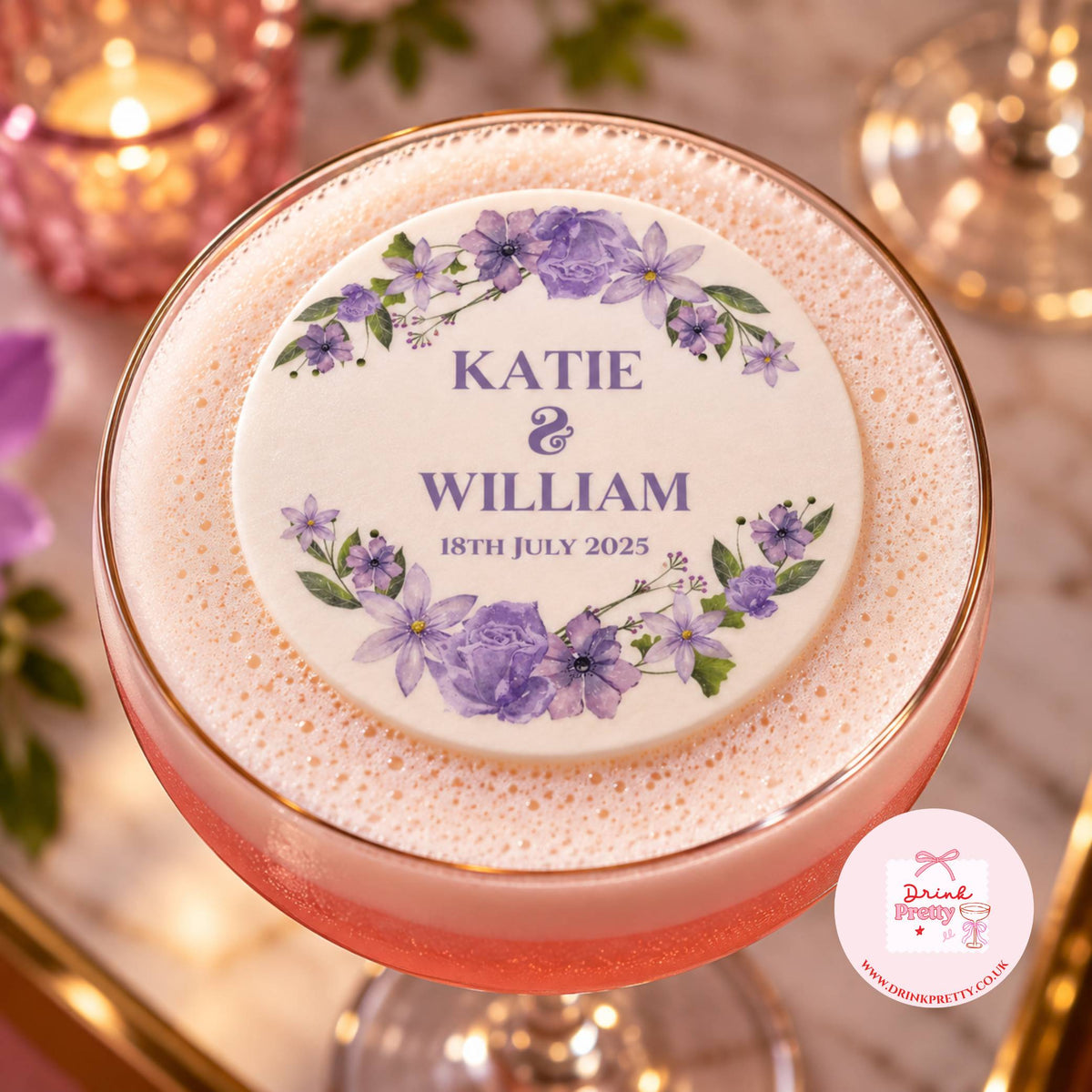 A close-up of a personalized round foam topper placed on top of a pink cocktail in a coupe glass. The topper is cream-colored with purple floral illustrations and the text 'KATIE & WILLIAM 18TH JULY 2025'.