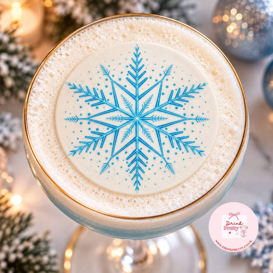A top-down view of a frothy beverage in a coupe glass with a gold rim, featuring a blue snowflake design on the creamy surface. The background is softly blurred with holiday lights and ornaments, and a small 'Drink Pretty' logo appears in the bottom right corner.