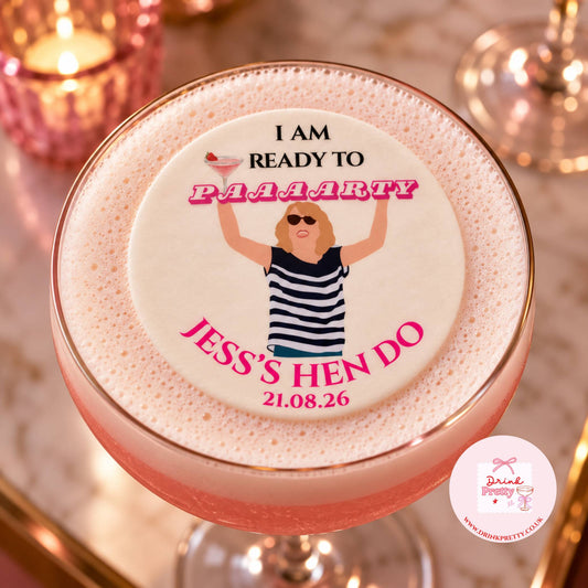 A pink cocktail in a coupe glass topped with thick white foam and a custom round foam topper. The topper features an illustration of a woman in a black-and-white striped top with the text 'I AM READY TO PAAARTY JESS'S HEN DO 21.