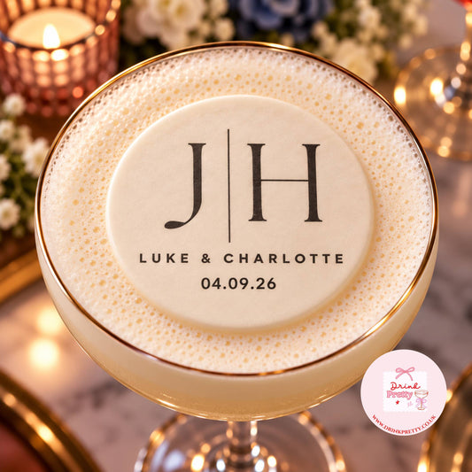 A personalized frothy cocktail in a coupe glass with a gold rim, topped with thick foam and a custom round label reading 'J | H LUKE & CHARLOTTE 04.09.