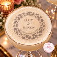 Load image into Gallery viewer, A close-up of a personalized cocktail in a gold-rimmed coupe glass, topped with thick foam featuring a custom leopard-print ring and the text 'LUCY & HENRY SATURDAY 19TH JULY 2020'. The background is softly blurred with warm candlelight and floral decor.
