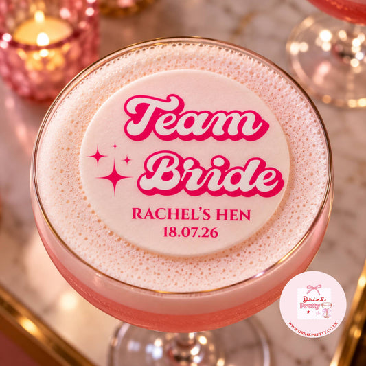 A pink frothy cocktail in a coupe glass with a round foam topper reading 'Team Bride Rachel's Hen 18.07.
