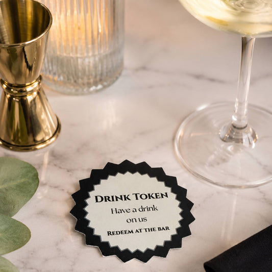 Black & white drink tokens Pack of 50 displayed on a table with a drink, offering a complimentary drink to guests.