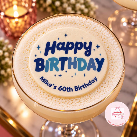 A close-up of a frothy beverage in a gold-rimmed glass, topped with foam printed with the words “Happy Birthday” and “Mike’s 60th Birthday” in blue and light blue lettering, with a small pink brand watermark in the lower right corner.