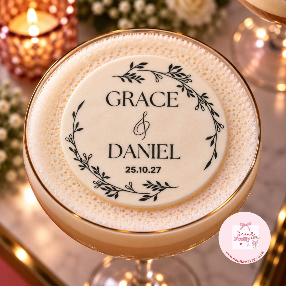 A close-up of a personalised round foam topper placed on top of a frothy cocktail in a coupe glass. The topper is cream-colored with black printed text reading 'GRACE & DANIEL' and a date, surrounded by a black leafy wreath design.