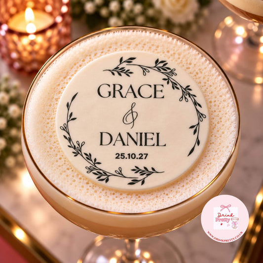 A close-up of a personalised round foam topper placed on top of a frothy cocktail in a coupe glass. The topper is cream-colored with black printed text reading 'GRACE & DANIEL' and a date, surrounded by a black leafy wreath design.