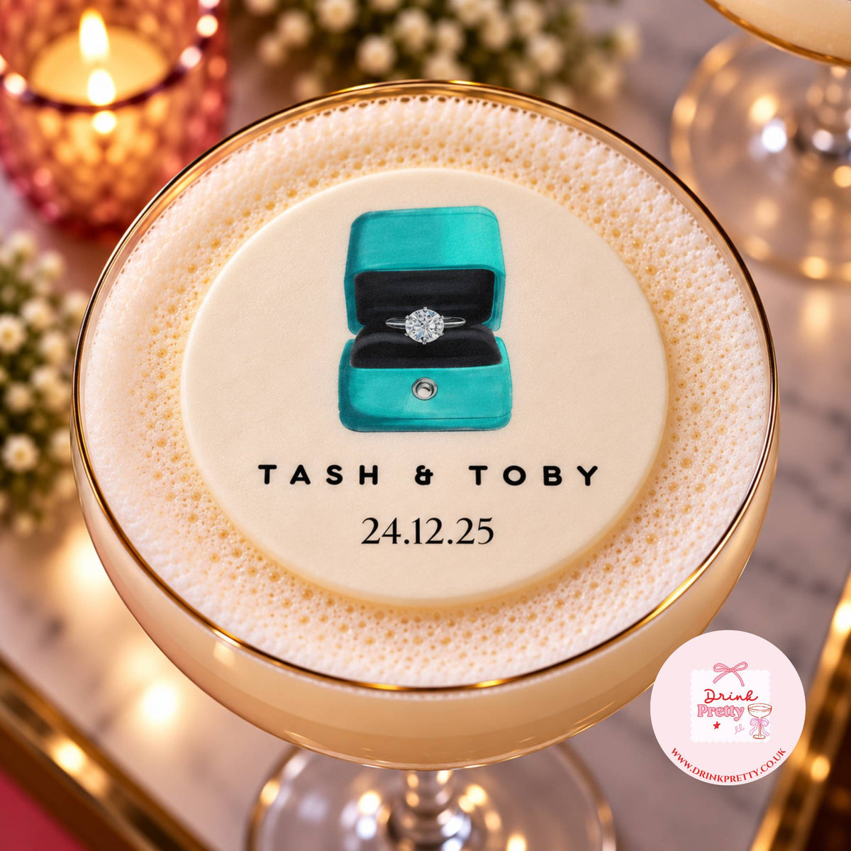 A close-up of a frothy cocktail in a coupe glass with a gold rim, topped with a custom illustration of a turquoise ring box and the text 'TASH & TOBY 24.12.