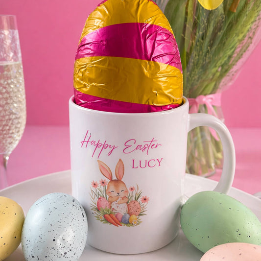 White mug with 'Happy Easter Lucy' design, filled with a large foil-wrapped Easter egg, on a pink background.