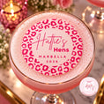 Load image into Gallery viewer, A pink cocktail in a coupe glass with a gold rim, topped with a thick white foam lid featuring a pink leopard print and the text 'Hattie's Hens MARBELLA 2026'. The background is a softly blurred, warm-toned bar or party setting with candles and glasses.
