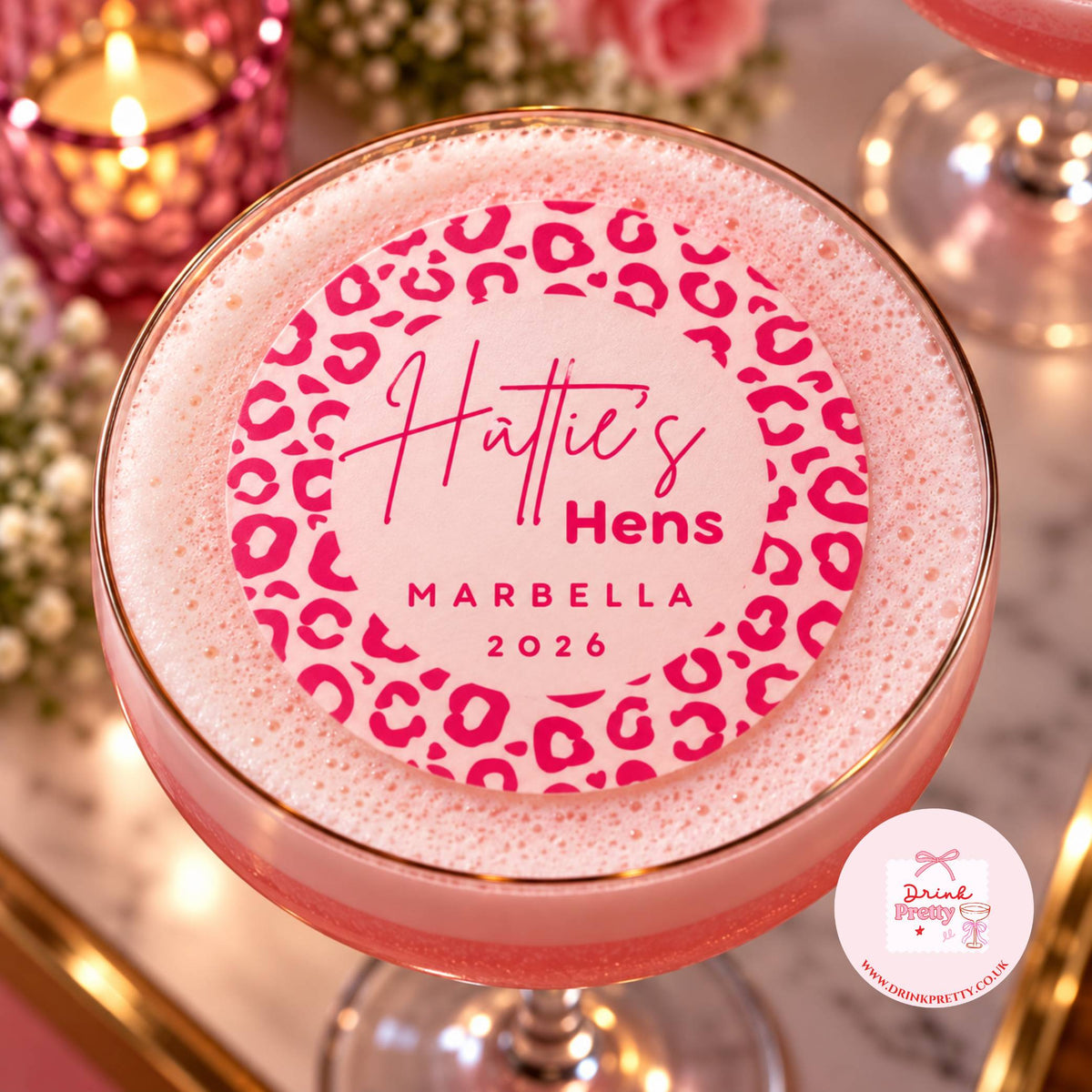 A pink cocktail in a coupe glass with a gold rim, topped with a thick white foam lid featuring a pink leopard print and the text 'Hattie's Hens MARBELLA 2026'. The background is a softly blurred, warm-toned bar or party setting with candles and glasses.