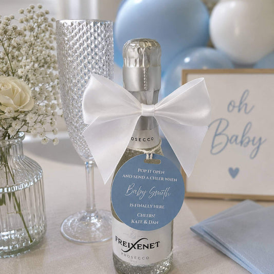 Baby shower name pop the fizz bottle favour tags on a prosecco bottle with a decorative bow and celebratory decor.