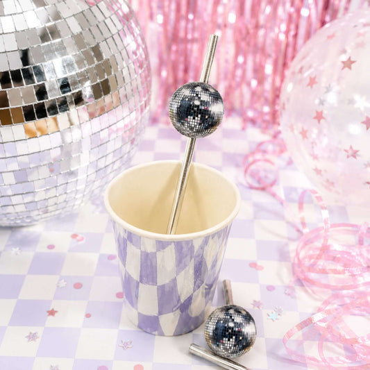 Disco ball paper party straw toppers in a cup surrounded by disco balls and party decorations.