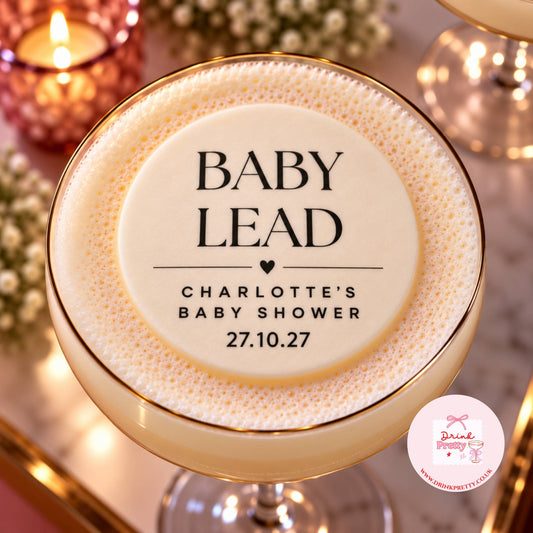 A close-up of a frothy champagne-colored cocktail in a coupe glass with a gold rim, topped with a printed foam label reading 'BABY LEAD' and 'Charlotte's Baby Shower 27.10.