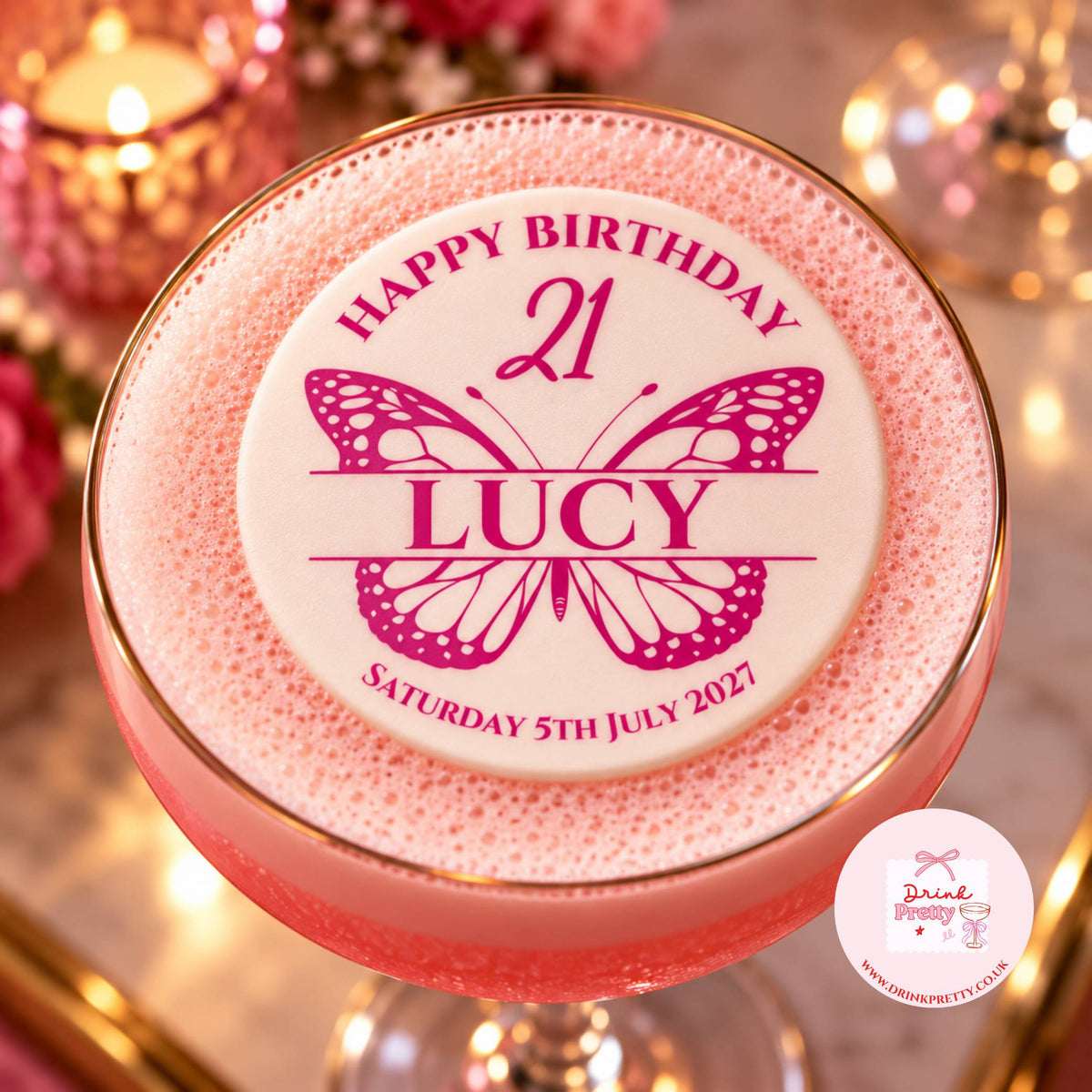 A pink frothy cocktail in a coupe glass with a gold rim, topped with a custom white foam lid that reads 'HAPPY BIRTHDAY 21 LUCY' with a butterfly design and the date 'SATURDAY 5TH JULY 2027'. The background is softly blurred with warm lights and pink floral decor.