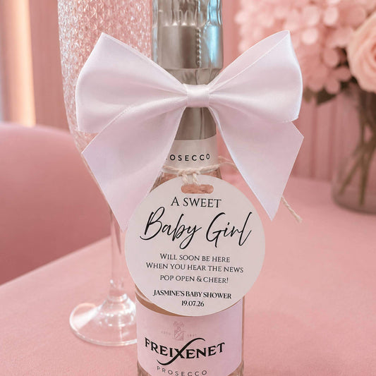Baby shower pop the fizz bottle favour tags on a prosecco bottle with a white bow and sweet announcement.