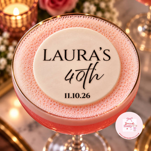 A close-up of a pink frothy cocktail in a coupe glass with a gold rim, topped by a round white topper printed with the text 'LAURA'S 40th 11.10.