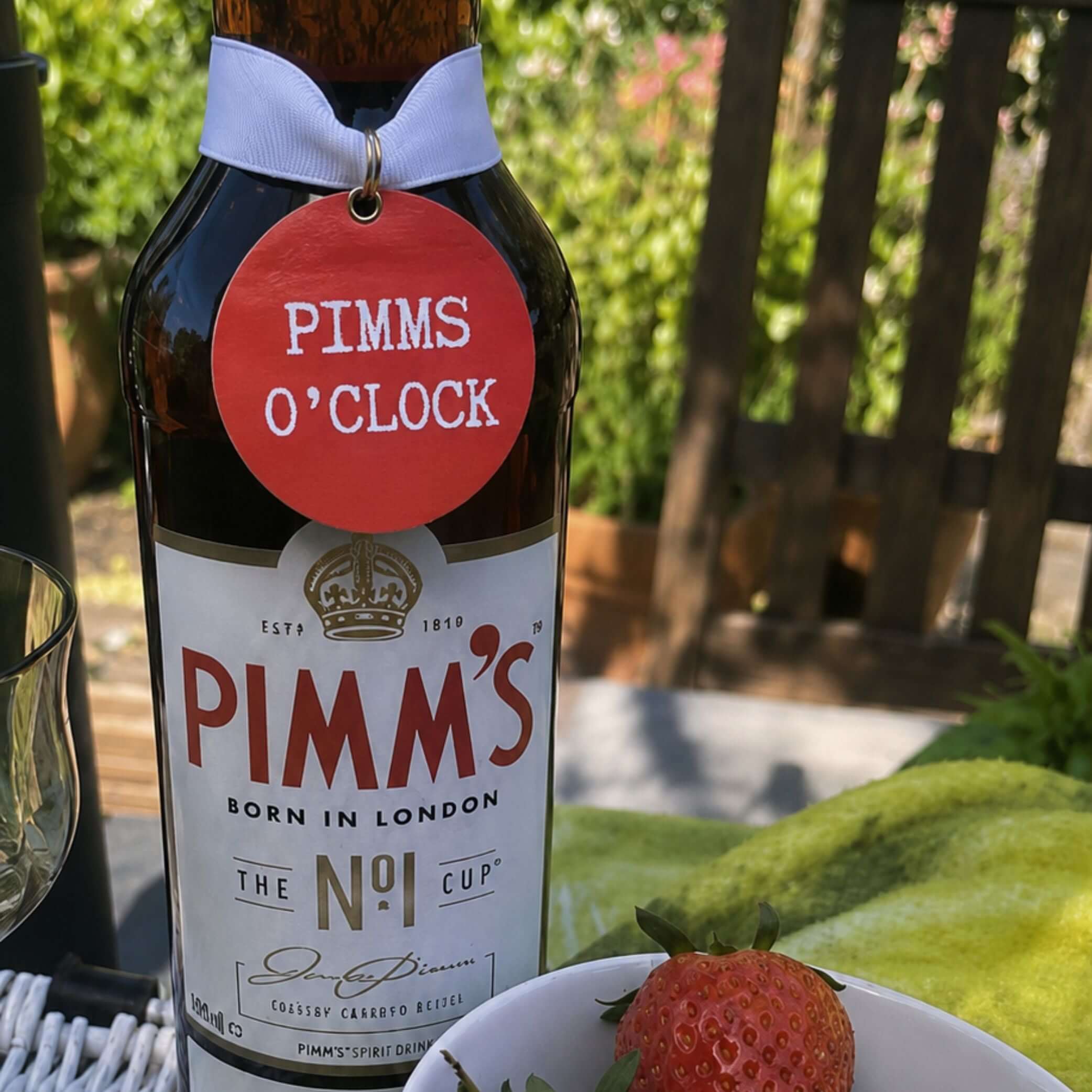 Pimms O'Clock Bottle Charm Gift Tag on a Pimm's bottle with fresh fruits in a garden setting.