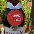 Load image into Gallery viewer, Pimms O'Clock Bottle Charm Gift Tag displayed on a Pimm's bottle in a sunny garden setting.
