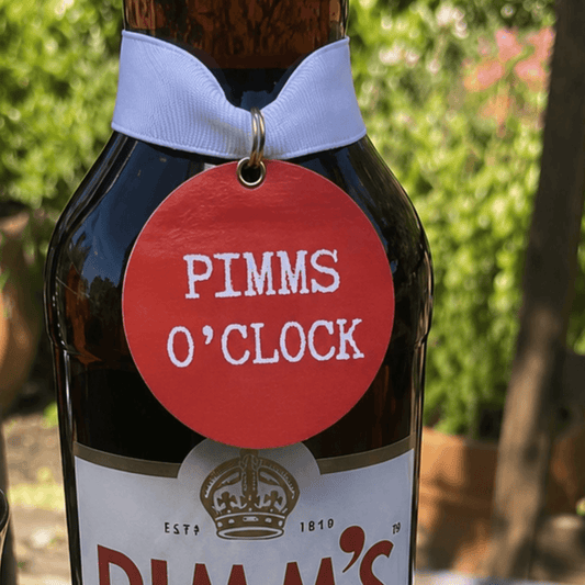 Pimms O'Clock Bottle Charm Gift Tag displayed on a Pimm's bottle in a sunny garden setting.