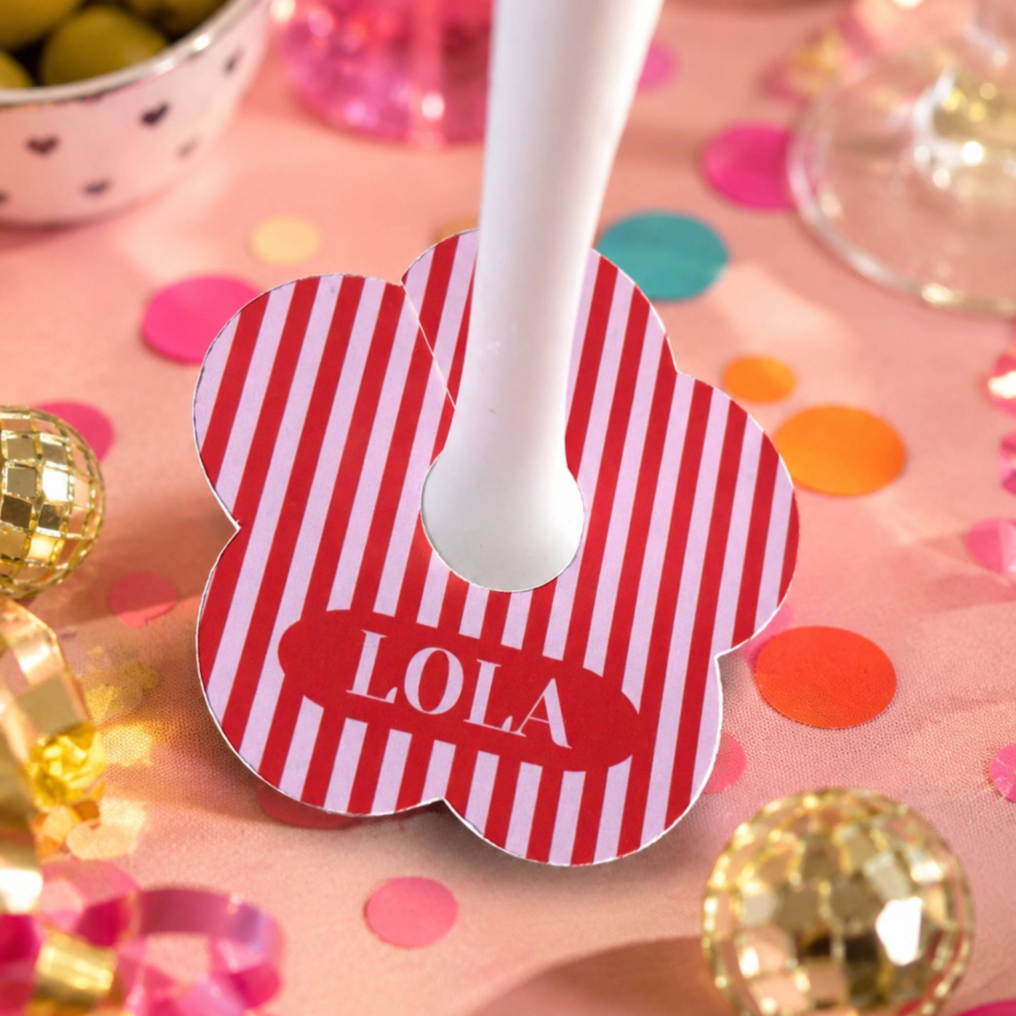 A personalised drink tag with red and light pink  vertical stripes, featuring a red oval label with the name 'LOLA' in white. A frosted glass is inserted into the centre of the tag, which is placed on a pink party table covered in confetti and party decorations.