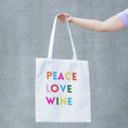 Load image into Gallery viewer, Peace Love Wine Tote Bag featuring colorful lettering on a white tote being held against a gray background.
