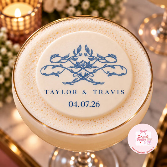 A top-down close-up of a frothy creamy beverage in a coupe glass with a gold rim, featuring a blue floral crest and the names 'TAYLOR & TRAVIS' with the date '04.07.