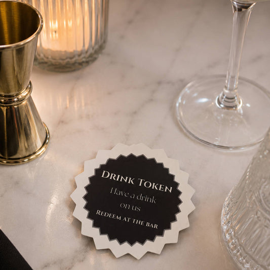 Black & white drink tokens Pack of 50 displayed on a marble table with bar tools, featuring text for redeeming drinks.
