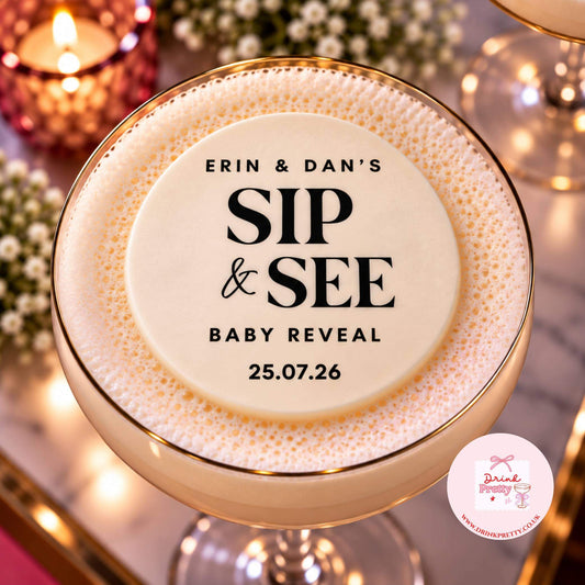 A close-up of a customised cocktail in a coupe glass, topped with thick foam and printed with the text 'ERIN & DAN'S SIP & SEE BABY REVEAL 25.07.