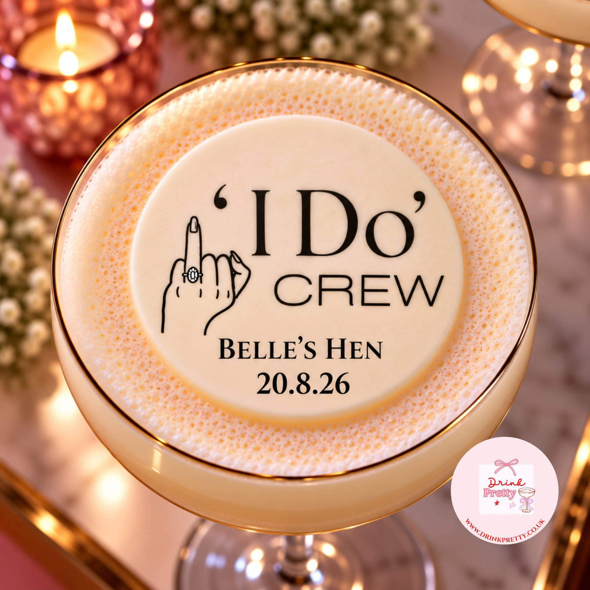 A close-up of a customised cocktail in a coupe glass with a gold rim, topped with thick foam featuring a custom printed design that reads "I Do Crew" and "Belle's Hen" with a date. The background is softly blurred with warm candlelight and floral decor.